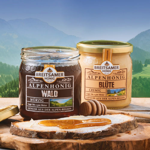 Breitsamer Alpenhonig Wald und Breitsamer Alpenhonig Blüte stehen vor einem Bergpanorama. Vor den Honiggläsern liegt eine Scheibe Brot mit Butter bestrichen und Honig auf einer Holzscheibe