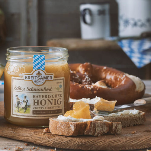 Holzbrett mit einem Glas Echtes Schmankerl Bayrischer Honig Blüte cremig, eine Scheibe Brot mit Butter und Honig und einer Brezel im Hintergrund