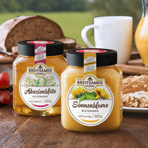 Breitsamer Sonnenblumenhonig und Breitsamer Akazienblüte, im Hintergrund ein gedeckter Frühstückstisch mit Brot, einer Tasse, und einer Karaffe Orangensaft