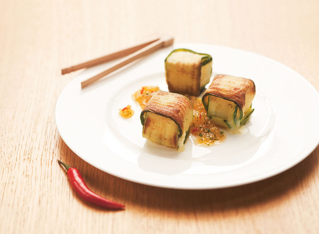 Drei Käse Päckchen mit Zucchini umwickelt auf einem weißem Teller. Vor dem Teller eine rote Chili Schote