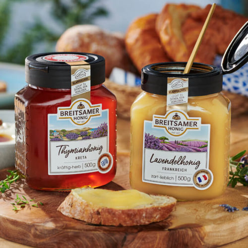 Breitsamer Thymianhonig aus Kreta und Breitsamer Lavendelhonig aus Frankreich auf einem Holzbrett mit einer Scheibe Brot mit Honig. Im Hintergrund ein Brotkorb mit Croissants