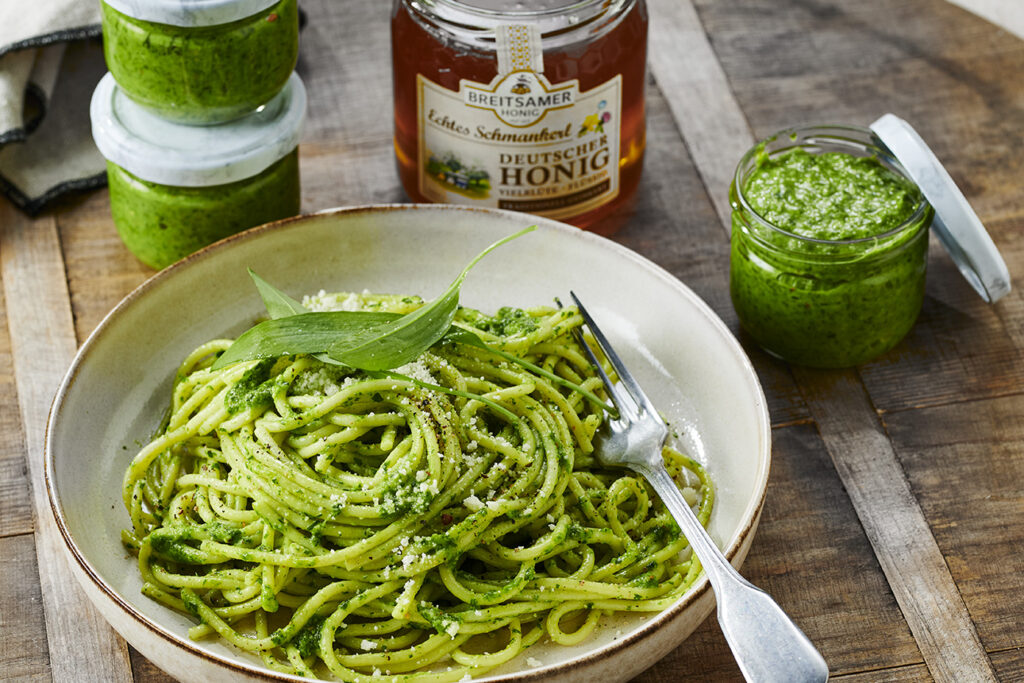 Teller mit Spaghetti und grünem Pesto. Im Hintergrund mehrere Gläser Honig-Bärlauch-Pesto und ein Glas Breitsamer Echtes Schmankerl Vielblüte.