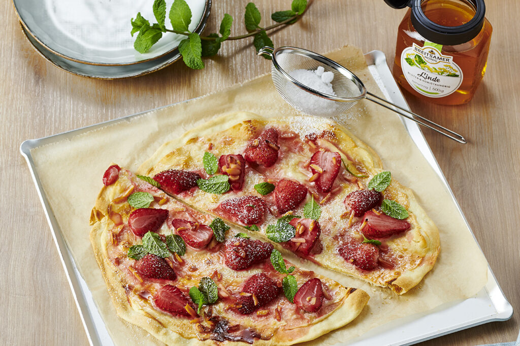 Backblech mit Backpapier, auf dem ein halbierter Flammkuchen, belegt mit Erdbeeren, liegt. Daneben ein Sieb mit etwas Puderzucker und ein Glas Breitsamer Lindenhonig