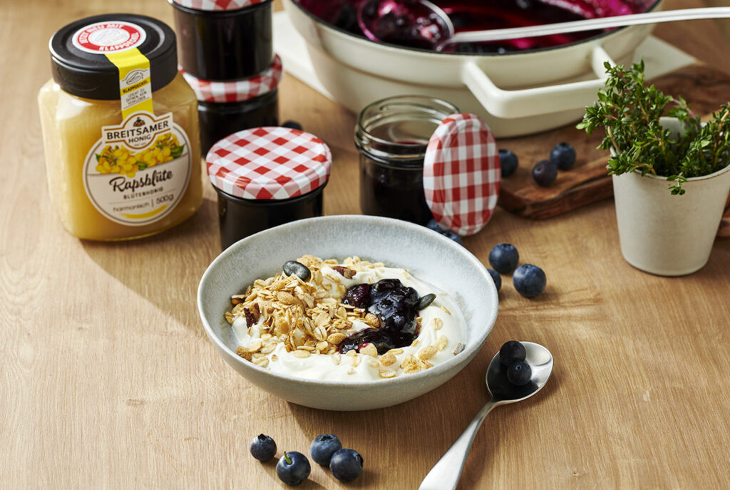 auf einem Tisch steht eine Schale mit Joghurt, Blaubeer-Honig-Konfitüre und Haferflocken. Dahinter stehen mehrere Gläser der Konfitüre sowie ein Glas Breitsamer Rapshonig. Vereinzelte frische Blaubeeren liegen auf dem Tisch